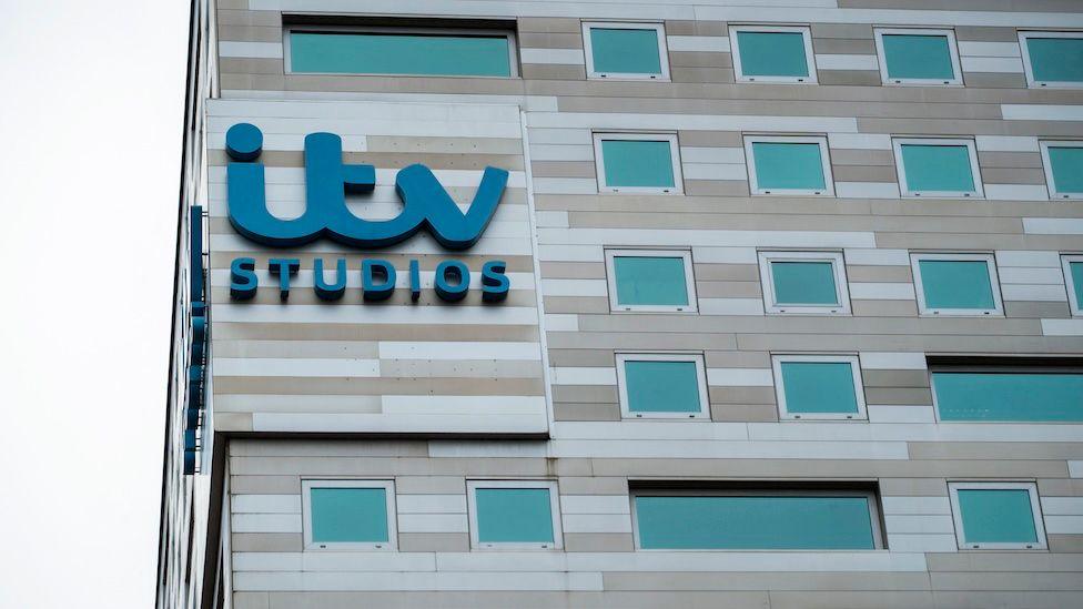The exterior of the ITV Studios office building in the Netherlands. The building itself is an uneven range of shades of light brown, with turquoise windows, and emblazoned with the company's logo in large turquoise letters.