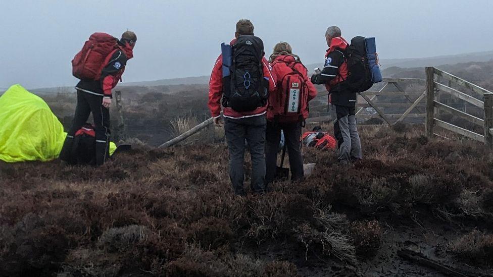 Four members of the rescue team standing on top of a very muddy fell on a grey drizzly day wearing backpacks and red coats and looking towards the ground where the man was stuck