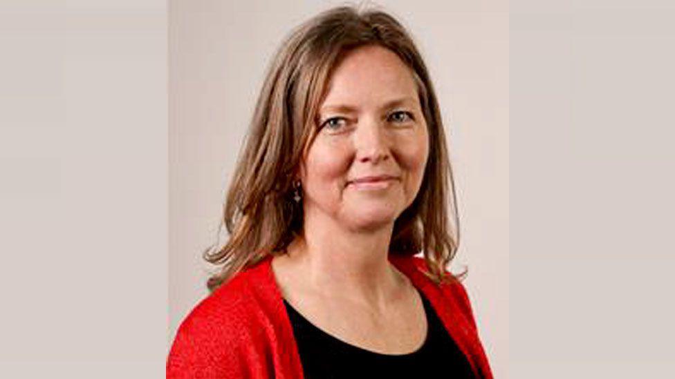 Mandy Clare is smiling. She has long brown hair and is wearing a black top and red cardigan.