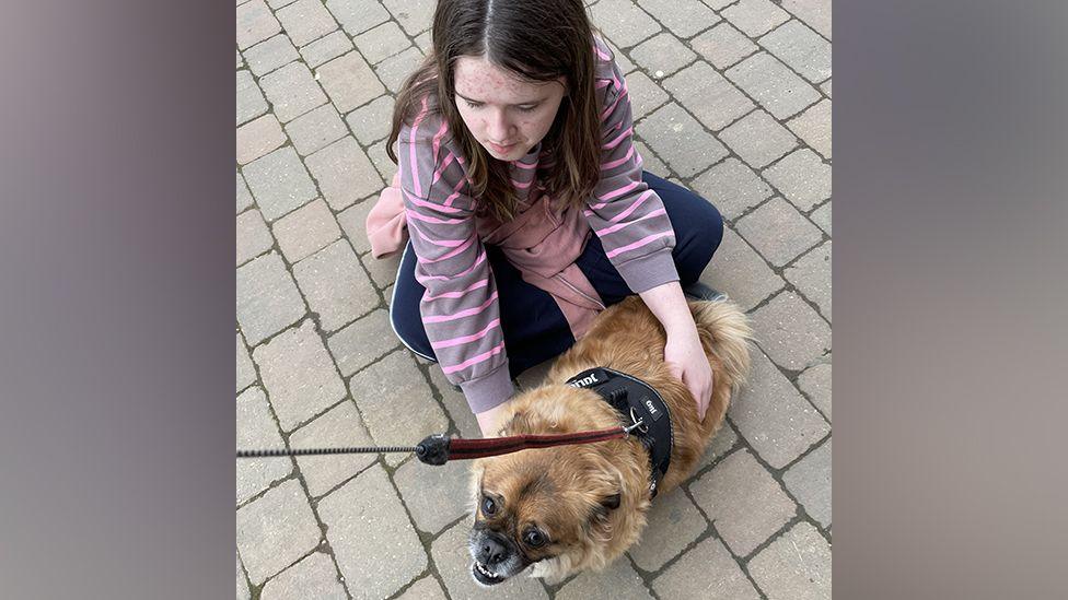 Rosie kneeling down holding a small dog, which is on a leash. Rosie is looking at the dog, with one hand on its back and another underneath. The dog is looking up and towards the camera and its teeth are just visible. Its lead is stretching up and out of frame, held by a hand that is not in shot. Both Rosie and the dog are on stone cobbles, which fill the rest of the frame.