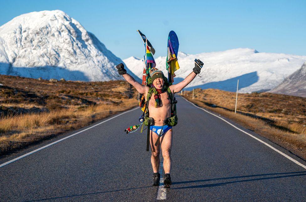 Mick Cullen, otherwise known as Speedo Mick, walks through Glencoe in sub-zero temperatures wearing only his swimming trunks. He is  wearing his blue Speedos, a woolly hat, scarf, backpack, gloves and nothing else, his face tilted upwards to the sky with his eyes closed and a look of relief, against a background of snowy mountains in Glencoe, Scotland, in January 2023