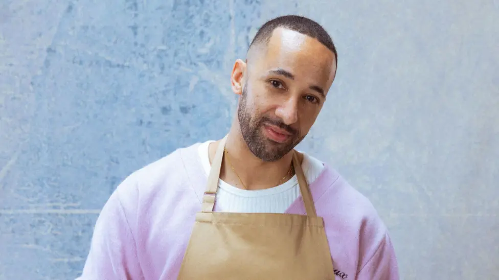 Aaron wearing a pink jumper and brown apron against a blue background