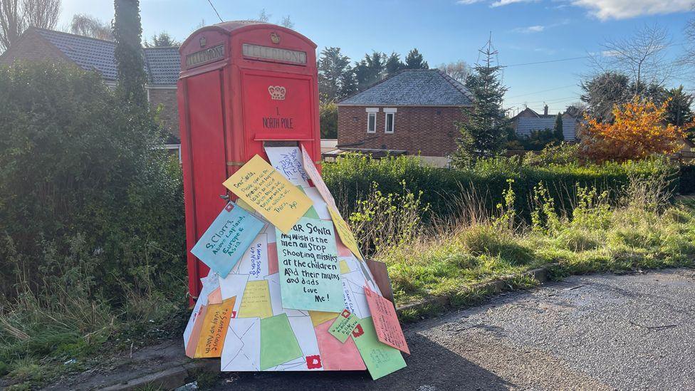 A phone box is made to look like a post box and there are lots of letters, cards and messages spilling out of it onto the ground
