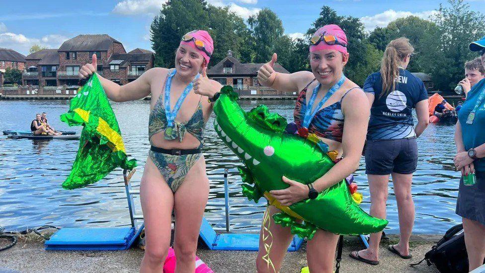 The twins in swimwear and pink swimming hats and goggles giving a thumbs up holding green metallic balloon crocodiles