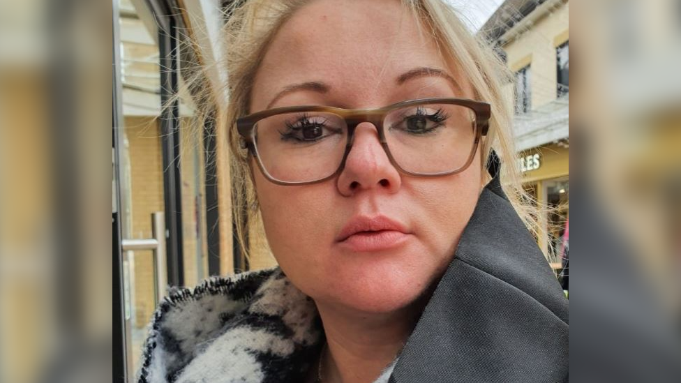 A head and shoulders shot of Gemma Whitehead. She has blonde hair tied back and is wearing brown glasses. She is wearing a grey coat and a black and white scarf. 