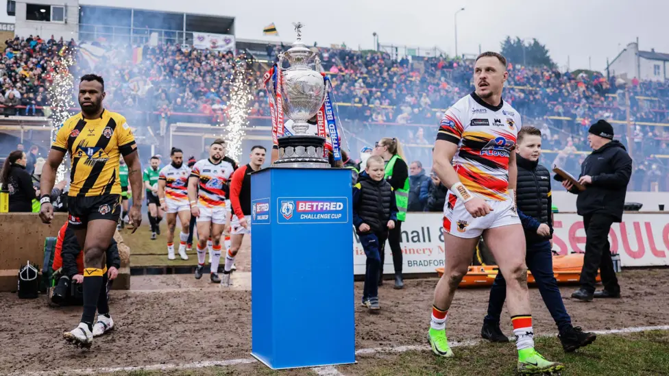 Players from Castleford and Bradford walk out of the tunnel at Odsal, across a shale motorsport track with wooden boards laid down to help avoid slipping, on to the grass pitch while the Challenge Cup stands on a plinth. Fireworks and smoke billow out as fans clap the players out of the dressing rooms.