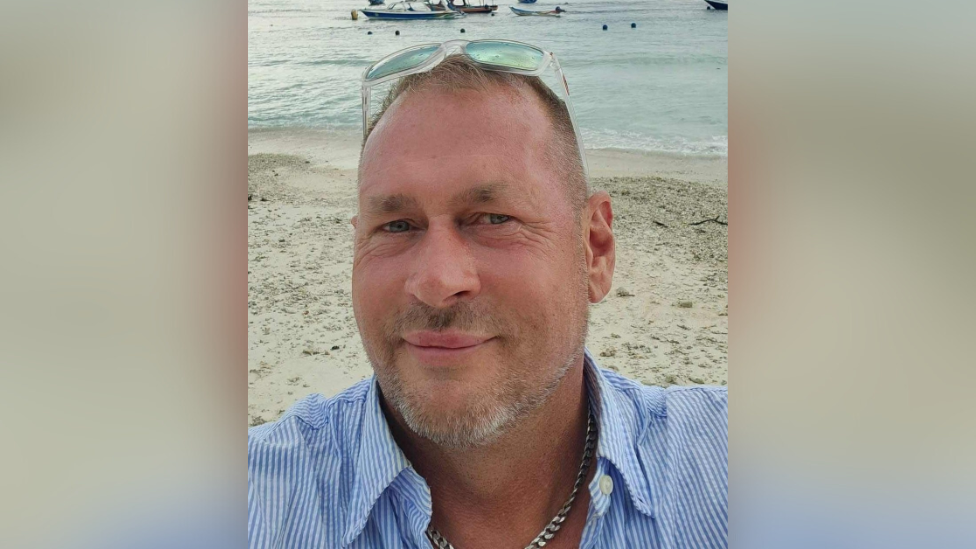 Robert Paczkowski. He has short brown hair and light beard. He is standing on the shore of a beach with water and boats in the background. He is wearing a light blue and white pin-stripe shirt and silver chain.