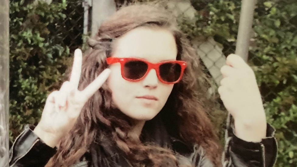 Maeve with red rimmed sunglasses on, her brown hair is partially tied back and she is doing a peace sign with one hand and holding what appears to be a swing with the other