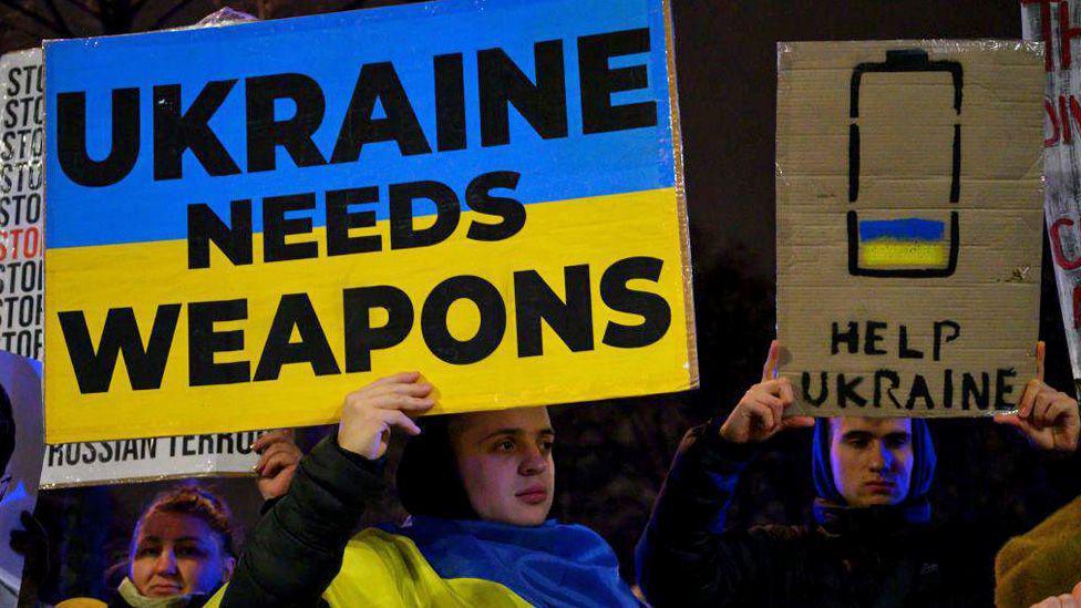 Demonstrators hold a signs at a protest outside the US embassy in Warsaw on 3 March, 2025. A man wearing a Ukrainian flag, hold a sign painted with the flag as the background and the words: Ukraine needs weapons, in black lettering. To the left of the man is another, holding a sigh with a nearly empty battery painted on it, filled slightly with the flag and the words Help Ukraine.