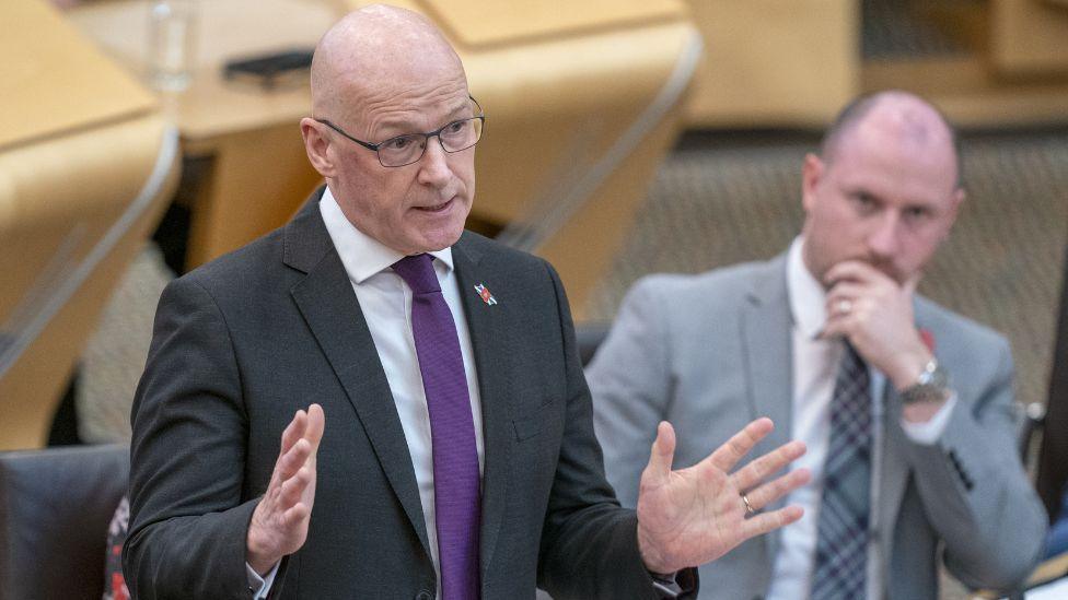 John Swinney, a bald man with a dark suit and purple tie, speaks in the Scottish Parliament with hands raised in front of him