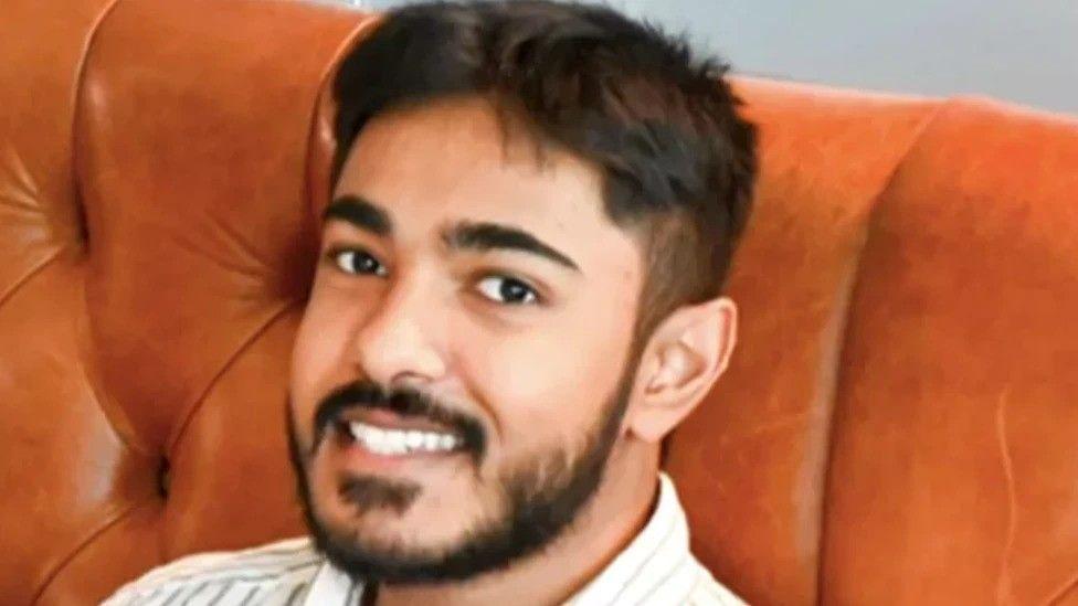 Anugrah Abraham, a man with black hair and facial hair, smiles into the camera. He is sat on brown leather sofa and the top of his shirt, which appears to be pinstriped, is visible.