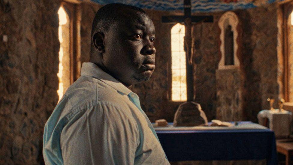 Ben Tekee as the character Shadrack, wearing a pale blue shirt, in the film Nawi. He is seen in a church - a giant cross seen on an small alter in the background