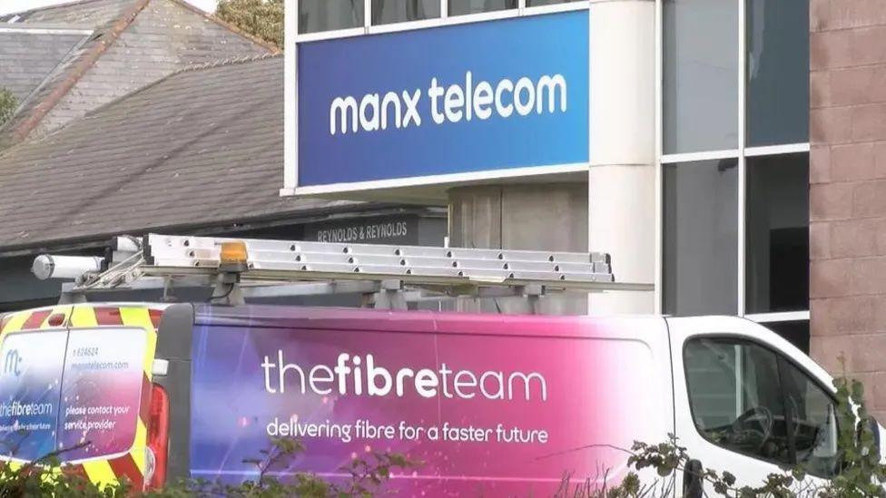 A van with a ladder lying flat on its roof with the slogan The fibre team, delivering fibre for a faster future on the side. It is parked outside a building that has a Manx telecom sign on the wall.