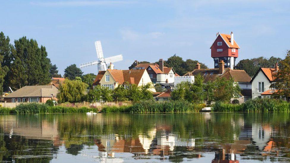 A boating lake in front of a village on a summer's day. The village is reflected in the water. A windmill can be seen in the distance behind some of the homes.