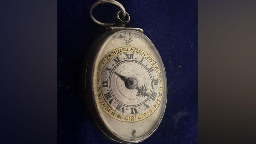 The front of a 17th Century silver oval-shaped pocket watch, with two rows of numerals, the inner Roman numerals from I to XII, the outer Arabic numerals from 1 to 31, the watch suspended from a loop at its top, against a black background