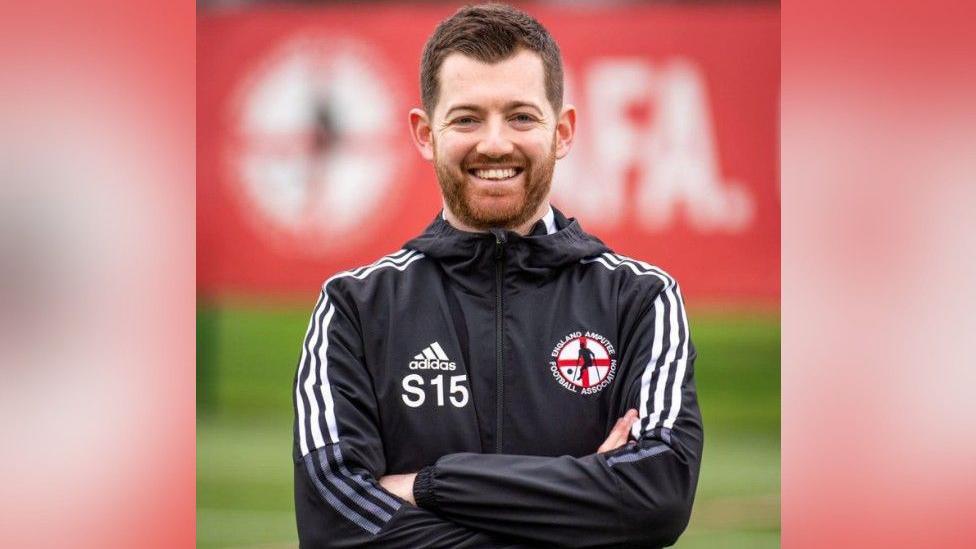 A man in a black jacket which says England Amputee Football Association, with a logo including an amputee kicking a ball in front of an England flag, smiles at the camera. His arms are crossed and the background is blurred.