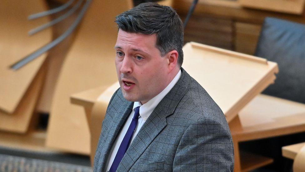 Jamie Hepburn, a dark-haired man wearing a patterned dark grey suit, speaks in the Scottish Parliament chamber. He is side on to the camera and visible from the chest up. 