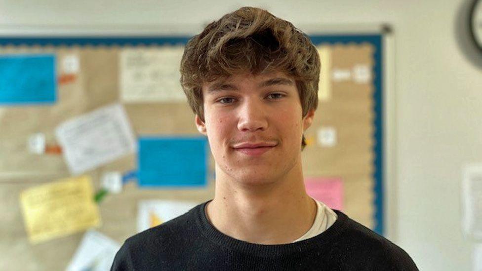 Charlie smiles at the camera, with a colourful noticeboard on the wall of the classroom behind him