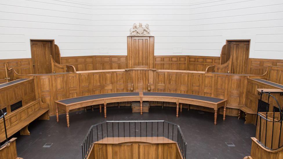 The inside of what used to be a court room at St Albans Museum. There is a wood panelled bench for judges or magistrates, a dock with railings in the middle of the room and a crest above a door.