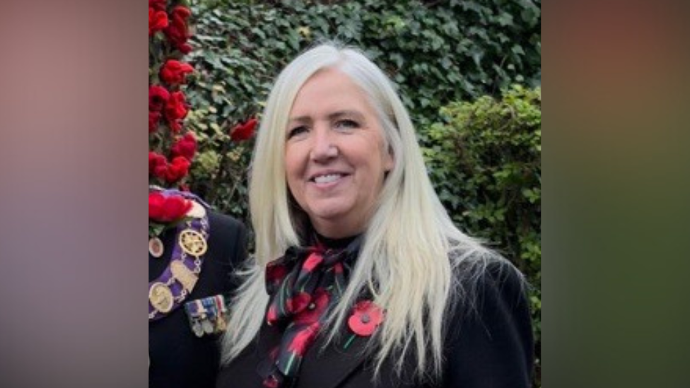 Headshot of blonde woman in a black jacket, wearing a poppy 