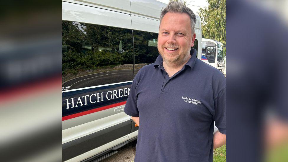 Nick is stood on the right-hand side of the picture, arms behind his back, smiling at the camera. He is stood next to one of his white minibuses, which says 'Hatch Green' on the side.