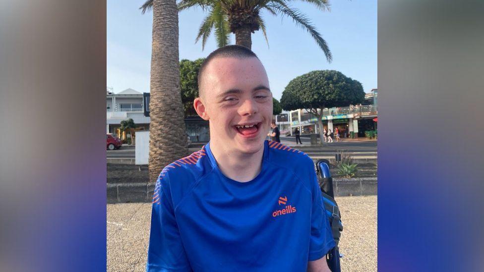 A young man with short brown hair, smiling at camera. He is sat in front of a palm tree and is wearing a blue O'Neill's top.