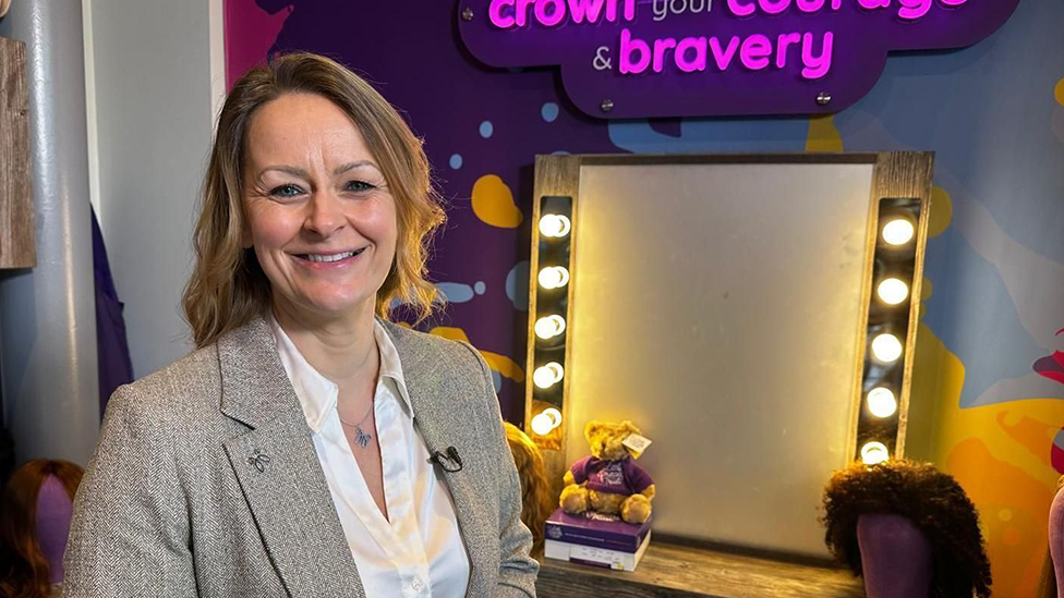 A woman with dark blonde hair down to her neck stands smiling in front of a theatre-style make-up mirror with lights down each side. She is wearing a white blouse and a grey jacket. A sign above the mirror reads: "These wigs are to crown your courage and bravery."