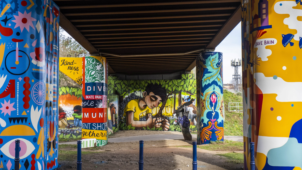 The underneath of a railway track bridge that has been covered in very bright and colourful murals and a large street art piece by Silent Hobo in the background. A grassy bank can be seen in the distance to the right.