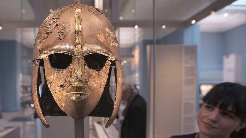 The Sutton Hoo helmet is a decorated Anglo-Saxon helmet found during a 1939 excavation of the Sutton Hoo ship-burial. It is facing the viewer and on the left while a woman is looking at it on the right.