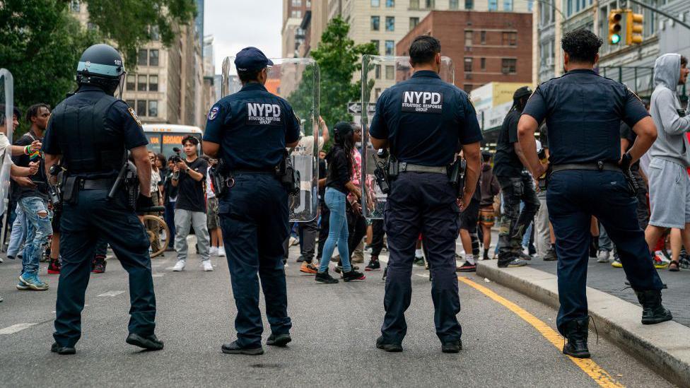 Four police officers have their backs to us as they provide security in Union Square as popular live streamer, not shown, stages a giveaway. They have NYPD on their shirts and one of them wears a helmet.