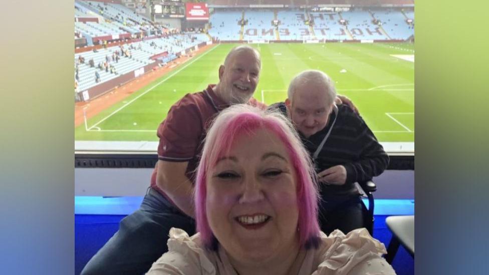 Anna-Marie has short pink hair and is smiling at the camera. In the background, a man is wearing a red shirt sitting next to Phil who is wearing a black and grey striped shirt. In the background is the Aston Villa football pitch 