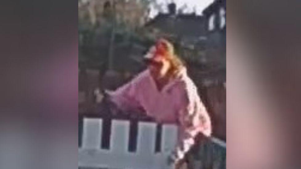 A grainy CCTV image of a woman in a pink hoodie and red baseball cap leaning over a white gate.