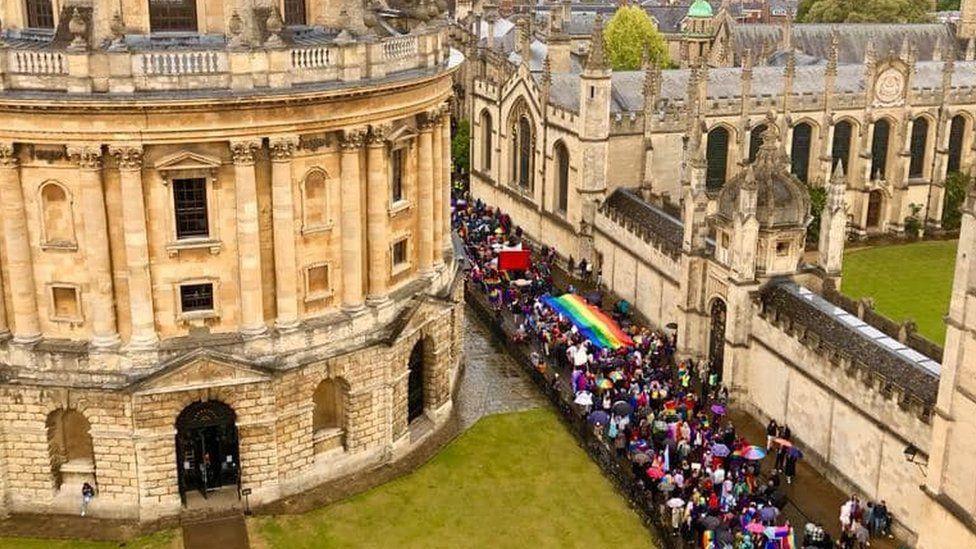 A stream of people walking past Oxford's Radcliffe Camera. They are colourful, and one section is holding up a large rainbow flag.