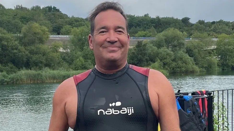 Mark O'Brien wearing black swimming vest standing in front of a lake, smiling