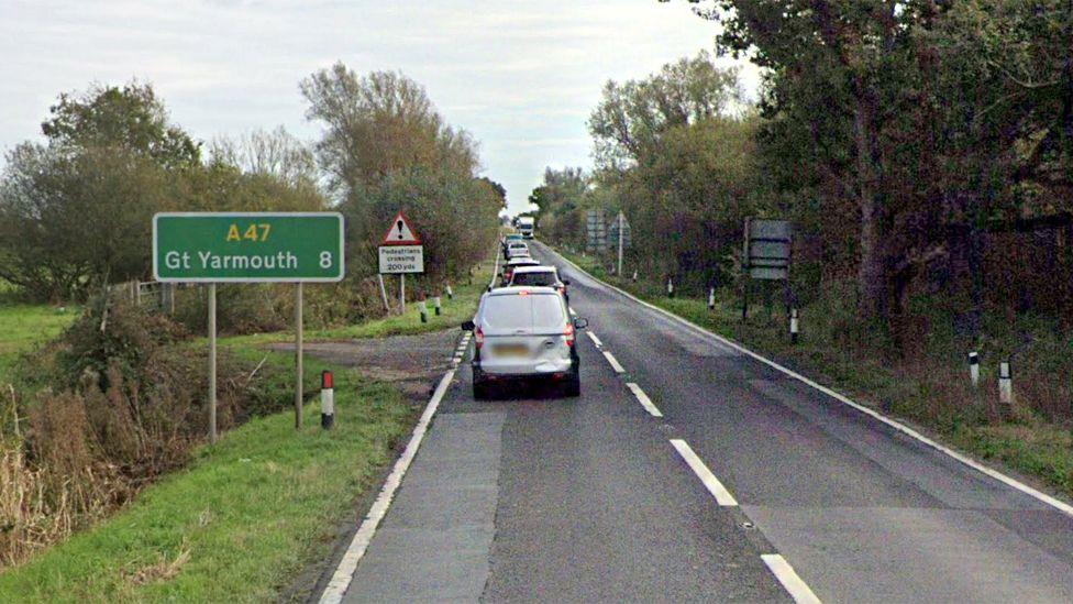 A stock image of the A47 Acle Straight showing cars backing up. The road is one lane in either directions. A green road sign showing Great Yarmouth is on the side of the road, which is surrounded by fields and shrubbery.