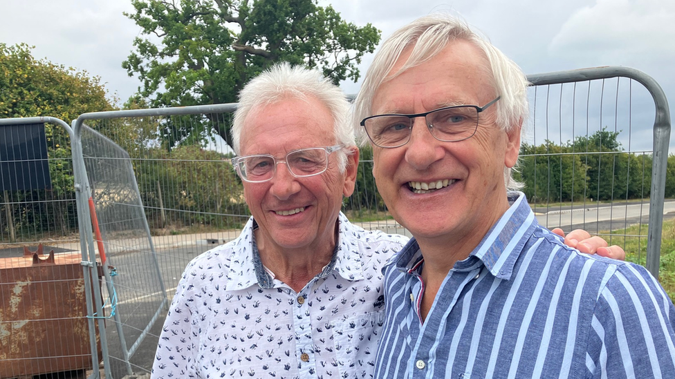 Two former male doctors wearing glasses smiling and standing in front of metal fencing 