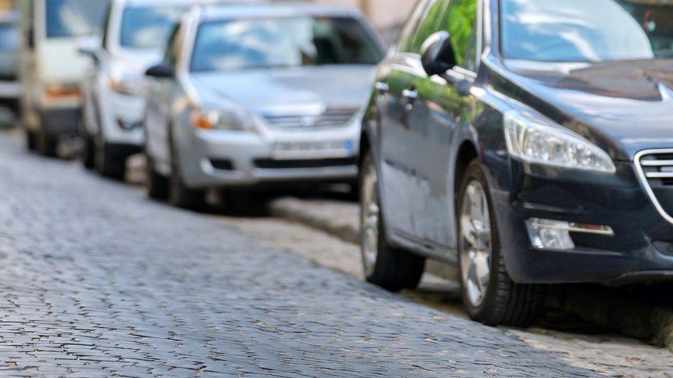 Cars parked half on the road and half on the pavement in a line one after another.