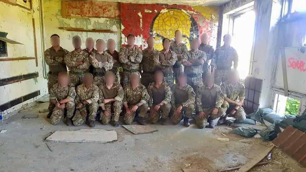 A group picture of Royal Marines in a dilapidated building. There is a row of marines stood up at the back and a row of marines kneeling in front of them. They are wearing camouflage fatigues.