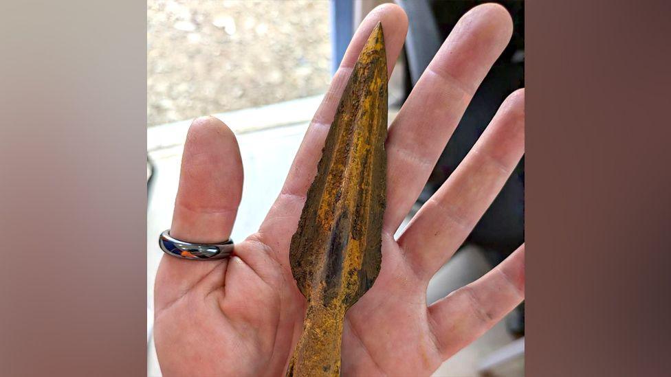 A medieval spearhead made of iron, held in a hand, which bears a ring on the thumb. The spear head has four fins, three of which are visible. It is showing signs of rusting, but is well preserved, and shaped to a point at the tip and curved towards the back end.