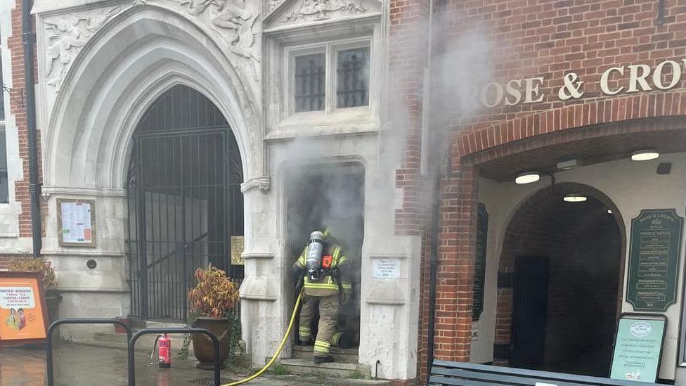 A firefighter entering the doorway of a property in Market Place, Saffron Walden, holding a hose, and wearing full breathing apparatus. There is a park bench to the right, two historic looking properties either side of the entrance. 