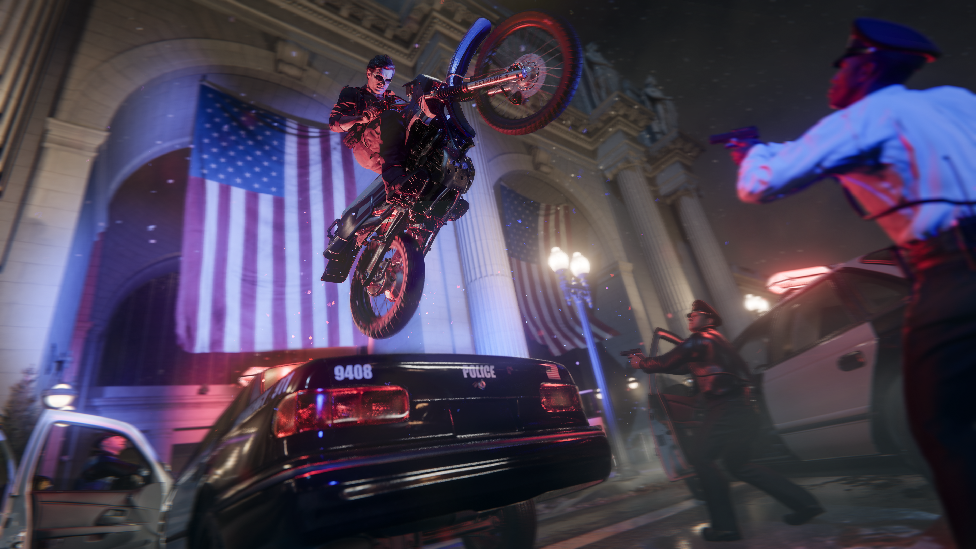 Screenshot shows a man in leather jacket and shades on a motorcycle sailing through the air over the top of a parked police car. On the ground two police officers with pistols drawn raise their weapons, looking up at the bike as it flies above them. In the background a white-fronted building with two very large American flags hanging inside large arches is visible.