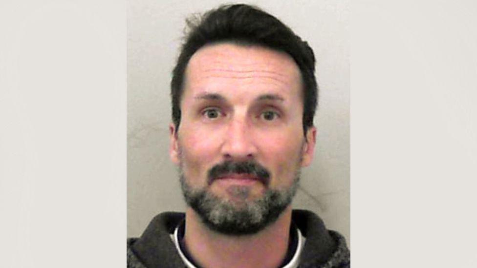 A police mugshot of Mark Acklom. He is wearing a white T-shirt underneath a black hoodie. He has short dark hair and a dark beard with grey patches. 