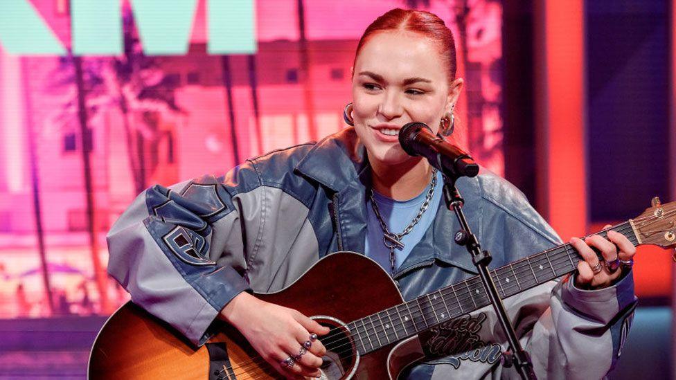 UPSAHL performing July 31, 2024 in Miami Beach, Florida. Shehas pulled back reddish hair and is wearing a grey and blue jacket, possibly leather. She is seated and playing a guitar, looking slightly over to her right, with a microphone on a stand in front of her