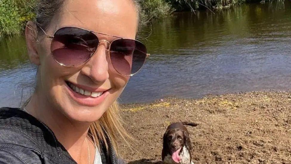 Nicola Bulley selfie smiling at the camera in sunglasses by the river with a brown and white spaniel by her side panting