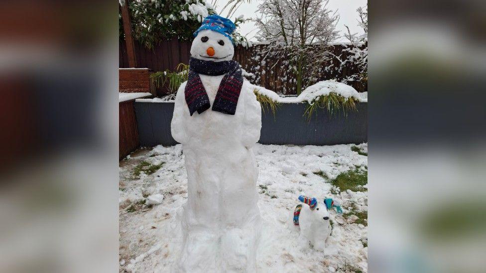 A snowman in a garden wearing a hat and scarf beside a small 'snow dog' in a garden. 