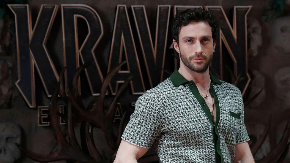 Aaron Taylor-Johnson posing in front of a large Kraven logo at a premiere