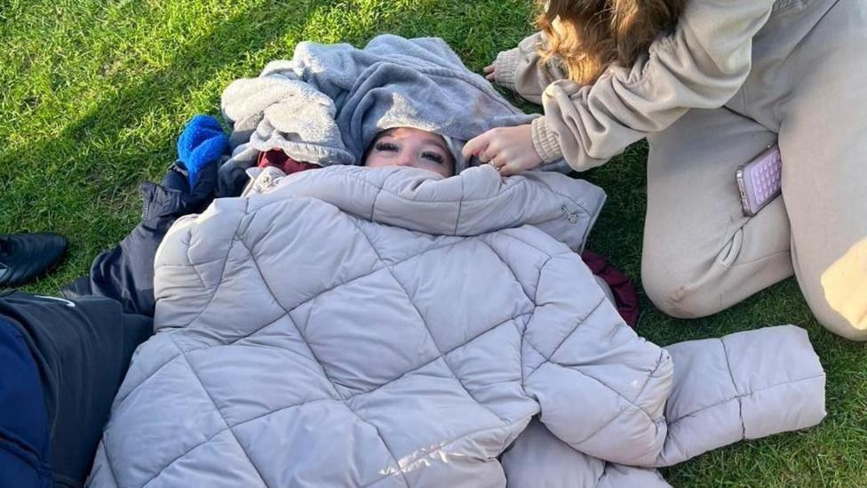 A girl lying on the ground, with jackets covering her. Other people can be seen sitting around her, trying to keep her warm.