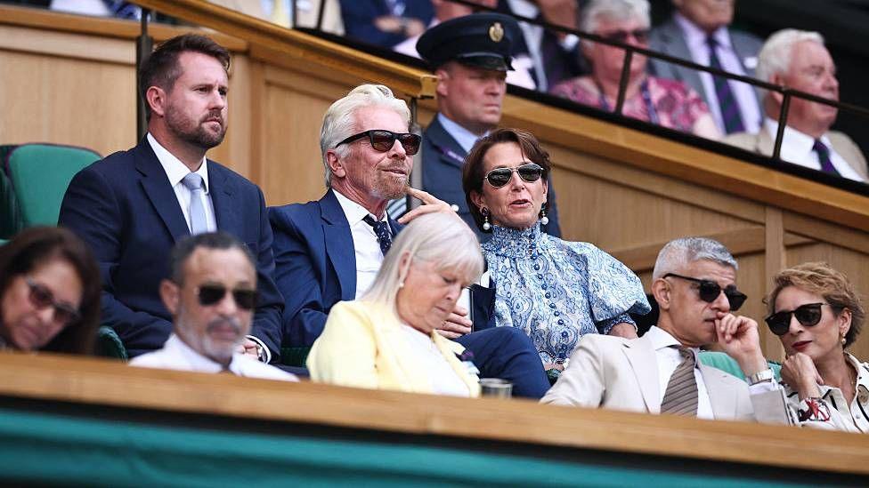 Richard Branson sits in the Royal Box on Centre Court alongside a woman wearing a blue dress and a man wearing a blue suit. He smiles as he watches the match.