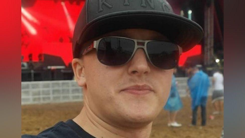 Selfie of Wojciech Trzebinski at a festival. He is wearing black sunglasses, a black t-shirt and a black baseball cap with KING embossed on it