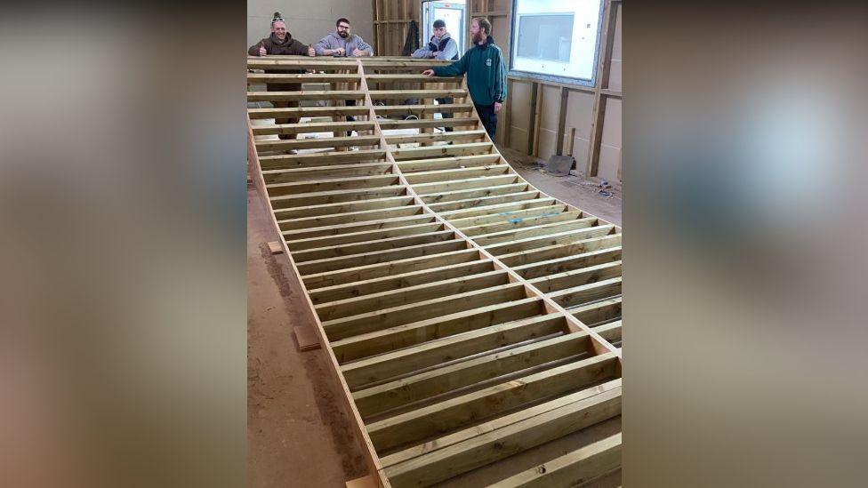 A group of four people stand at the end of a skateboarding ramp which is being constructed. It is a large wooden structure with the slats visible - the skating surface has not yet been added.